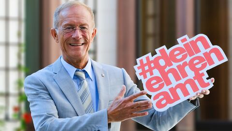 CdS #deinehrenamt Staatsminister Wintermeyer hält das Logo #deinehrenamt hoch und lächelt in die Kamera.