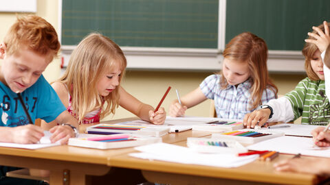 Kinder sitzen an einem Tisch vor einer grünen Tafel im Klassenraum und arbeiten an ihren Aufgaben