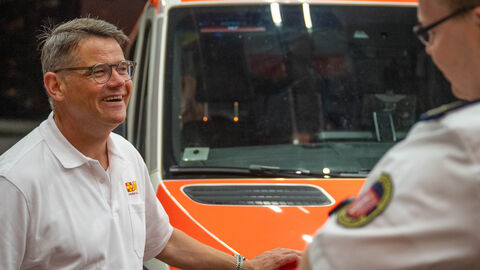 Ministerpräsident Rhein begleitet den ASB Ministerpräsident Rhein vor einem Einsatzfahrzeug im Gespräch mit einer weiteren Person.