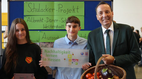 Bildtext: Schulacker Projekt. Nachhaltigkeit lernen in Hessen. Umweltstaatsekretär Oliver Conz und zwei Schüler lächeln in die Kamera. Übergabe der "Umweltschule".