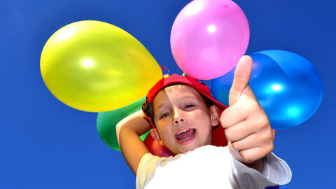 Daumen hoch! Kind steht mit bunten Luftballons vor dem blauen Himmel und streckt den Daumen nach oben