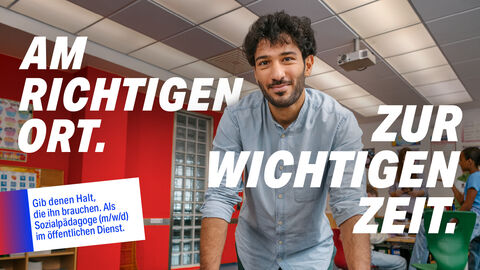 Am richtigen Ort zur wichtigen Zeit_Sozialpädagoge Ein Sozialpädagoge steht im Klassenzimmer. Auf dem Bild steht der Text: "Am richtigen Ort zur wichtigen Zeit. Gib denen Halt, die ihn brauchen. Als Sozialpädagoge (m/w/d) im öffentlichen Dienst."