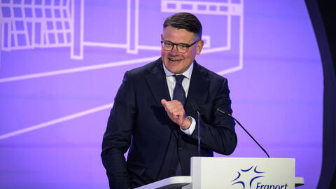 Ein Mann steht vor einer lilafarbenen Wand mit weißen Linien und spricht