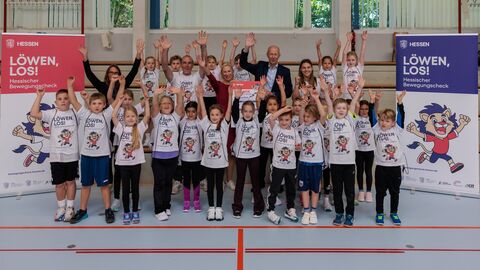 Gruppenfoto in einer Turnhalle mit Grundschulkindern und Erwachsenen, die jubelnd die Arme heben; links und rechts stehen Banner mit der Aufschrift „LÖWEN, LOS! Hessischer Bewegungscheck“.
