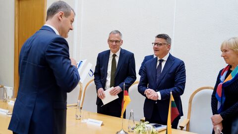 : Ministerpräsident Boris Rhein und litauischer Regierungschef Gintautas Paluckas Zwei Männer stehen vor einem Tisch. Dahinter steht ein weiterer Mann, der zu ihnen spricht und ein Geschenk in der Hand hält.