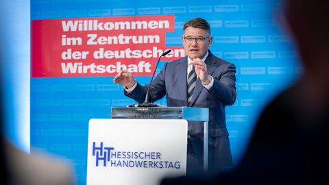 Ministerpräsident Boris Rhein steht an einem Rednerpult. Hinter ihm steht eine blaue Wand mit einem roten Balken und weißer Schrift "Willkommen im Zentrum der deutschen Wirtschaft"